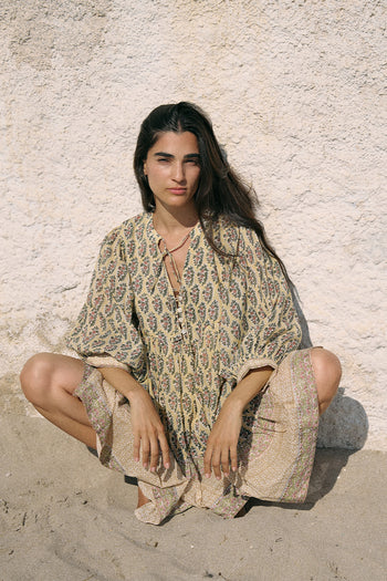 Woman sitting cross-legged on sand wearing the Daughters of India Bhoomi Mini Dress in Meadow, a sage green and cream paisley block print cotton mini dress with billowed sleeves