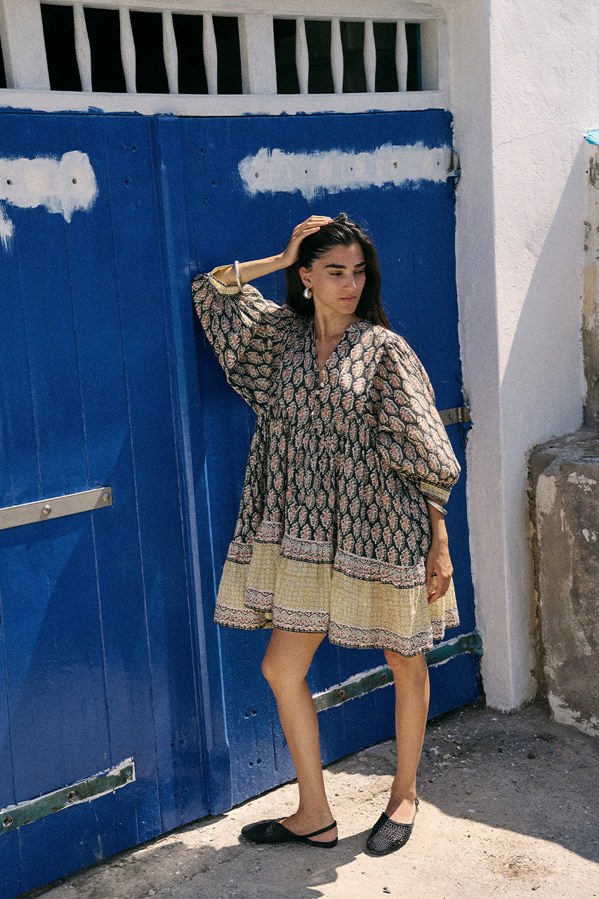 Full-length view of woman in front of blue double doors wearing the Daughters of India Bhoomi Mini Dress in Onyx, showing the complete mini dress silhouette and flat shoes