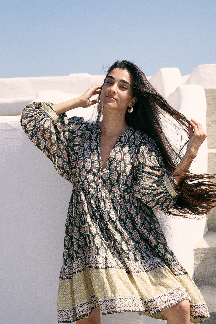 Woman with windswept hair against a white Mediterranean wall wearing the Daughters of India Bhoomi Mini Dress in Onyx with billowed sleeves and tiered skirt