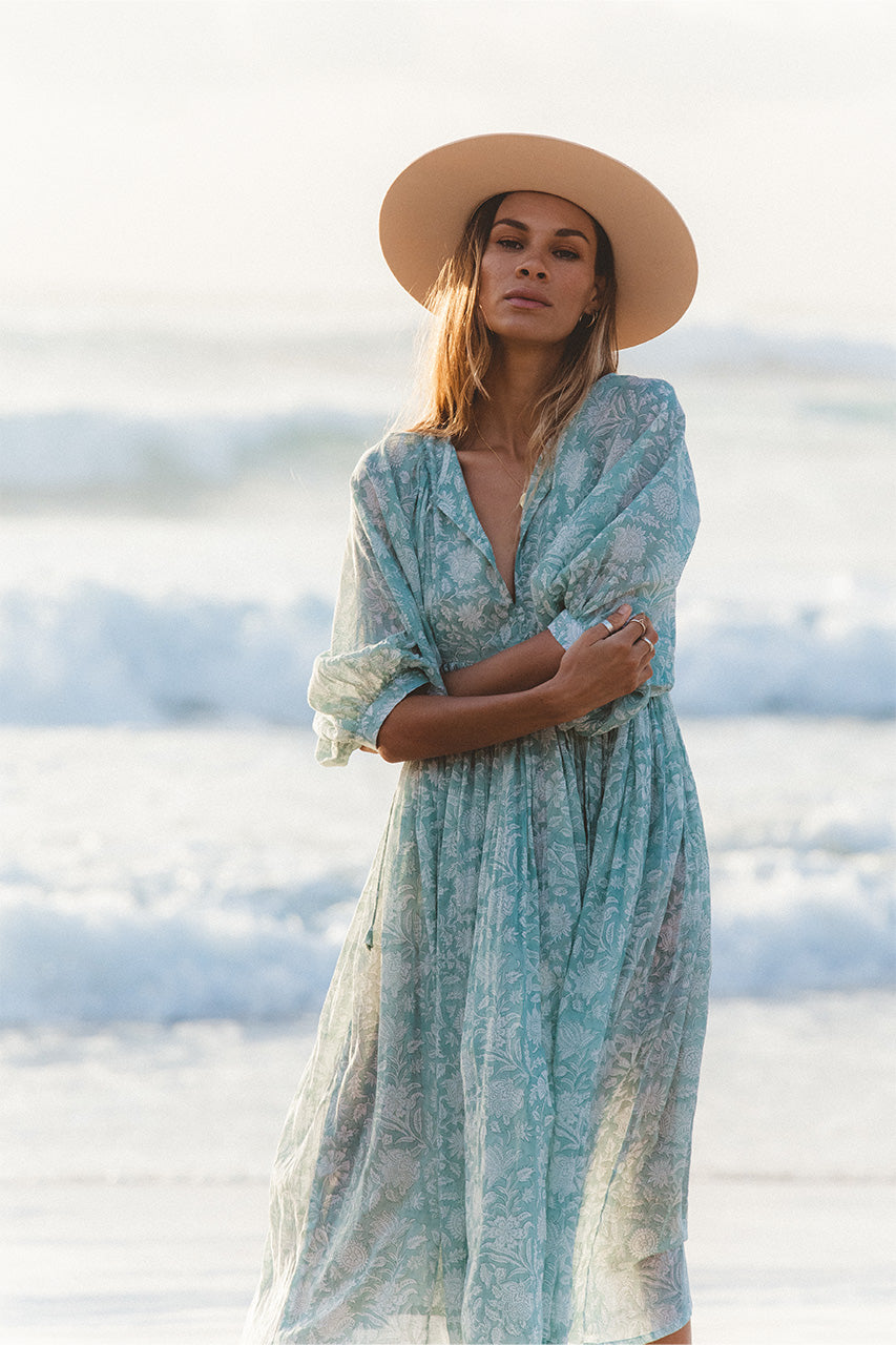 Woman wearing Daughters of India Kyra Midi Dress in Jade with a wide-brim sun hat, standing on a beach with ocean waves in the background