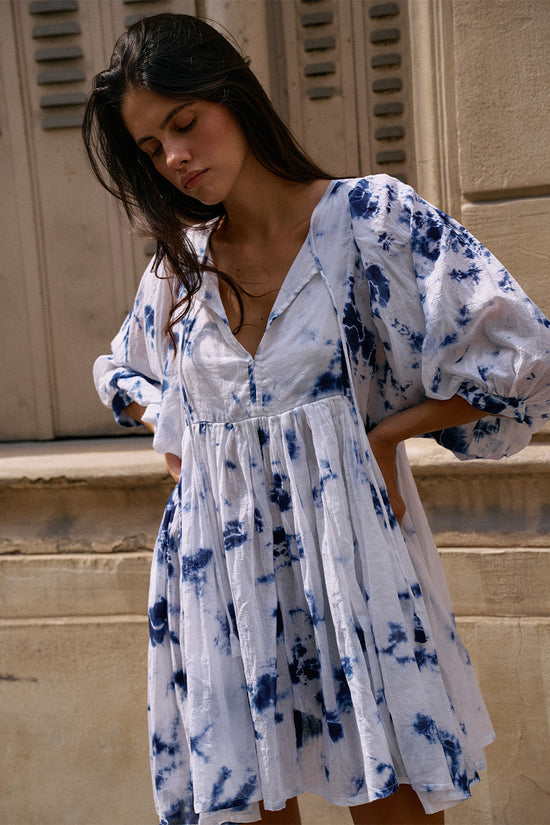 Kyra Mini Dress Deep Sea — close-up front view at stone building entrance, deep sea shibori tie-dye cotton mini dress with V-neckline and balloon sleeves, European shuttered facade | Daughters of India